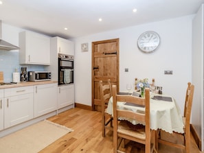 A compact kitchen with white cabinets, a microwave, and an oven. There's a wooden door, a dining table with chairs, and a wall clock. - Fox Bank Cottage (Macclesfield)