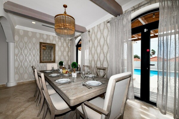 A dining area with a wooden table set for four, white upholstered chairs, a chandelier, patterned wallpaper, and a view of a swimming pool through a glass door.
