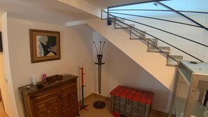 A staircase with a glass railing, a wooden cabinet with drawers, a floor lamp, and a bench with a red and gray patterned cover.
