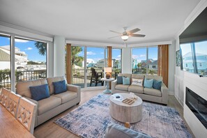 A spacious living room with a large window, a ceiling fan, and a fireplace. - Indies 101 | Modern Corner Beach Condo | Spacious Balcony Retreat (Gulf Shores)