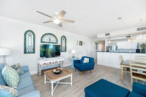 Ein modernes Wohnzimmer mit einer Couch, einem Couchtisch und einem Fernseher. Es gibt einen Essbereich mit Tisch und St ühlen. Im Hintergrund ist eine Küche mit weißen Schränken und einer Arbeitsplatte zu sehen.