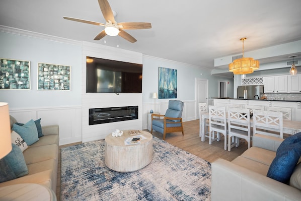 A modern living room with a ceiling fan, a flat-screen TV, and a dining area with a kitchen in the background. - Indies 101 | Modern Corner Beach Condo | Spacious Balcony Retreat (Gulf Shores)