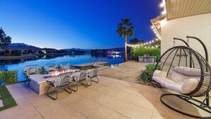 A patio with a fire pit, outdoor seating, a hanging chair, and a view of a swimming pool at dusk. - The Lakefront Social: An Extraordinary Waterfront Experience (Indio)