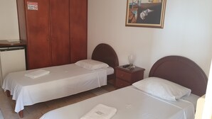 Habitación de hotel con dos camas individuales, un armario de madera, una mesita de noche con una lámpara y un cuadro en la pared.