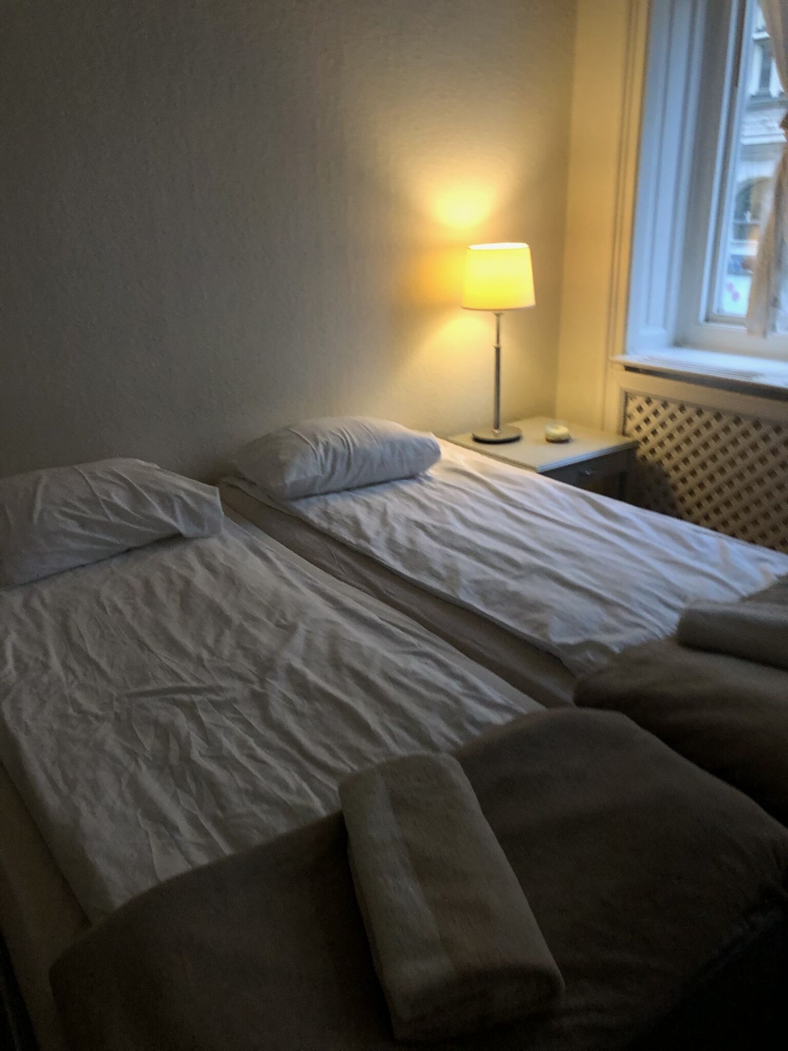 A hotel room with two beds, a nightstand, a lamp, and a window with a view of buildings.