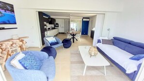 Un salon moderne avec un canapé, des fauteuils et une table basse. On y trouve également un coin repas avec une table et des chaises, et une cuisine à l’arrière-plan.