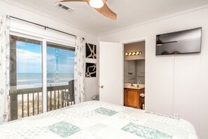 A bedroom with a bed, a television, a bathroom with a mirror and sink, and a view of the beach through a sliding door.
