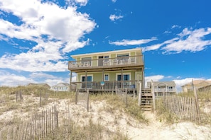 Ein Strandhaus mit zwei Etagen, einem Balkon, umgeben von Dünen und spärlicher Vegetation.