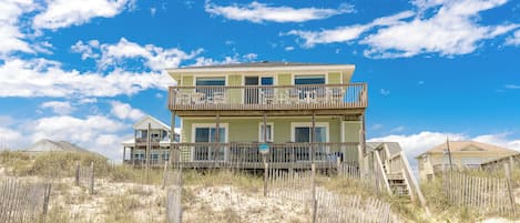 A two-story beachfront property with a balcony on the upper level, a sandy beach in the foreground, and a clear blue sky.