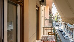 A balcony with a table and chairs, a view of the street, and an air conditioning unit. - 2br renovated flat close to metro (Athens)