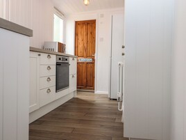 Una cocina con armarios blancos, horno empotrado y piso de madera. Hay un letrero que indica un baño.