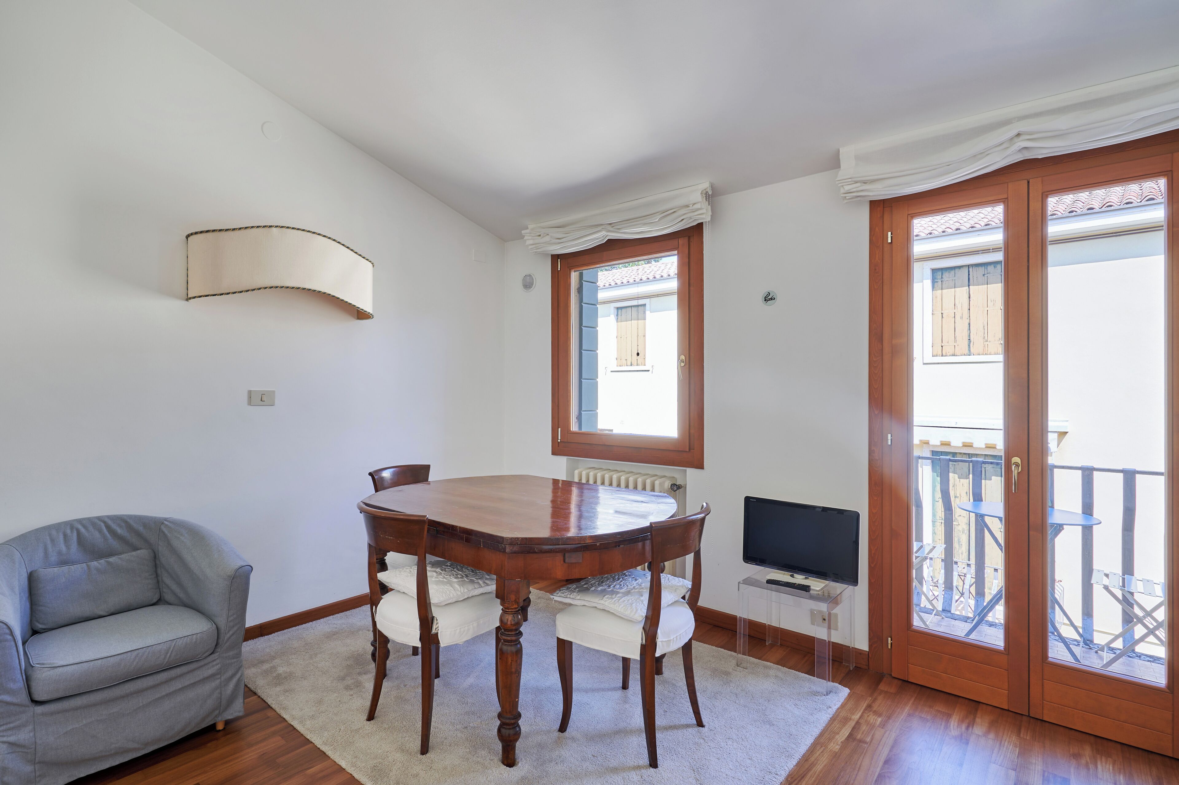 A dining area with a wooden table and chairs, a grey sofa, a TV on a stand, and a balcony with a wooden railing.