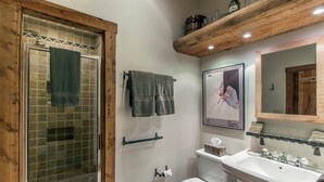 Un baño con cabina de ducha de vidrio, inodoro, lavamanos y estante de madera sobre el lavamanos.