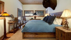 Un dormitorio con una cama grande, cabecera de madera, dos ventanas, una lámpara y una cabeza de búfalo montada en la pared.