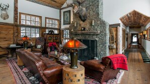 Interior rústico de una cabaña con sofás de cuero, una chimenea de piedra y un ciervo montado en la pared.