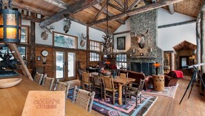 Interior rústico de una cabaña con una mesa de madera, sillas, chimenea y cornamentas de ciervo decorando las paredes.