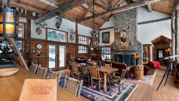 Interior rústico de una cabaña con una mesa de madera, sillas, chimenea y cornamentas de ciervo decorando las paredes.