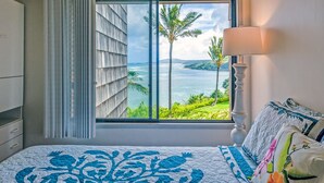A bedroom with a large window offering a view of the ocean and palm trees. - Oceanfront views all the way to the lighthouse! (Princeville)