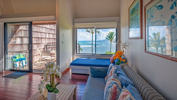 A room with a large window overlooking the ocean, a bed with blue bedding, a wicker sofa, and a small table with flowers. - Oceanfront views all the way to the lighthouse! (Princeville)