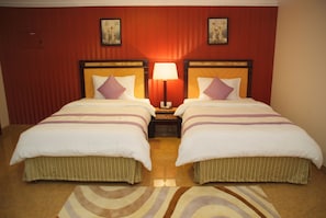 A hotel room with two beds, a nightstand with a lamp, and a patterned rug. - Garden Hotel Muscat (Muscat)
