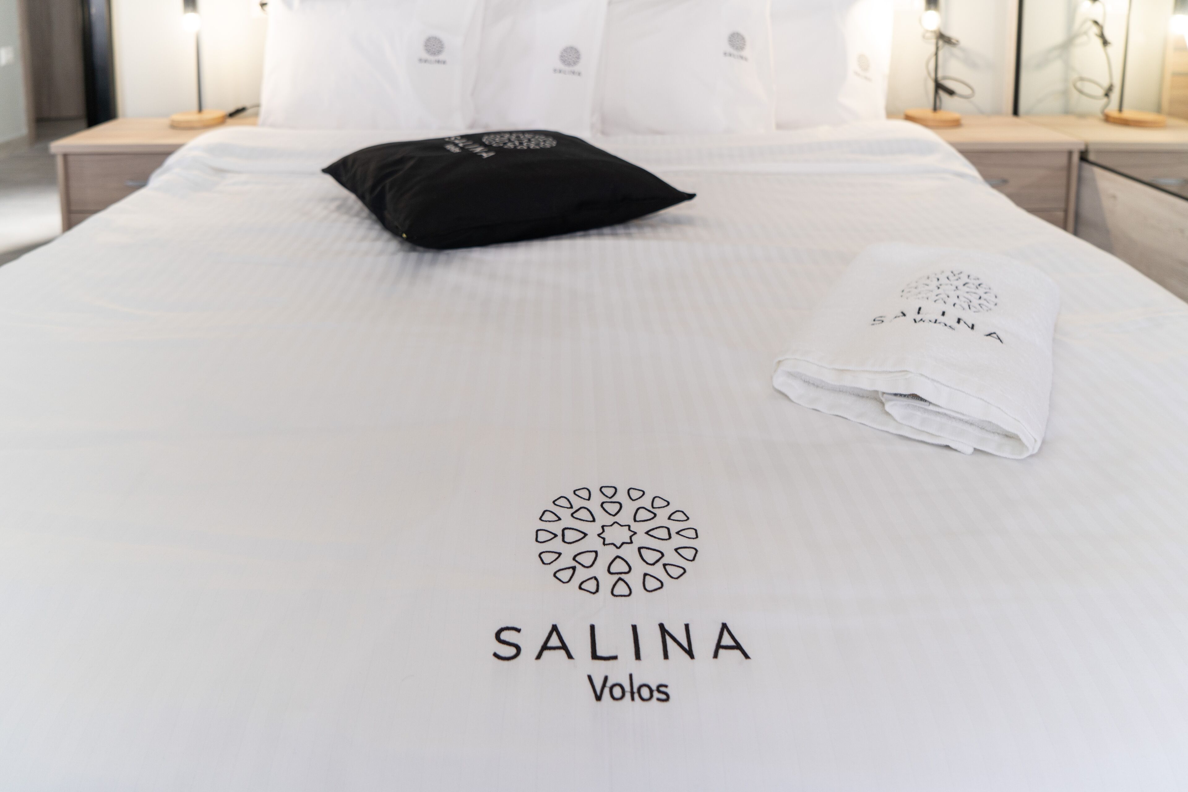 Een net opgemaakt bed met een witte spreidek die een logo en de naam 'SALINA Volos' in het midden heeft.