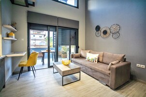 Una sala de estar moderna con un sofá, una mesa de centro y una silla. Se ve una ventana con persianas, un estante con objetos decorativos y una pared con detalles artísticos circulares.