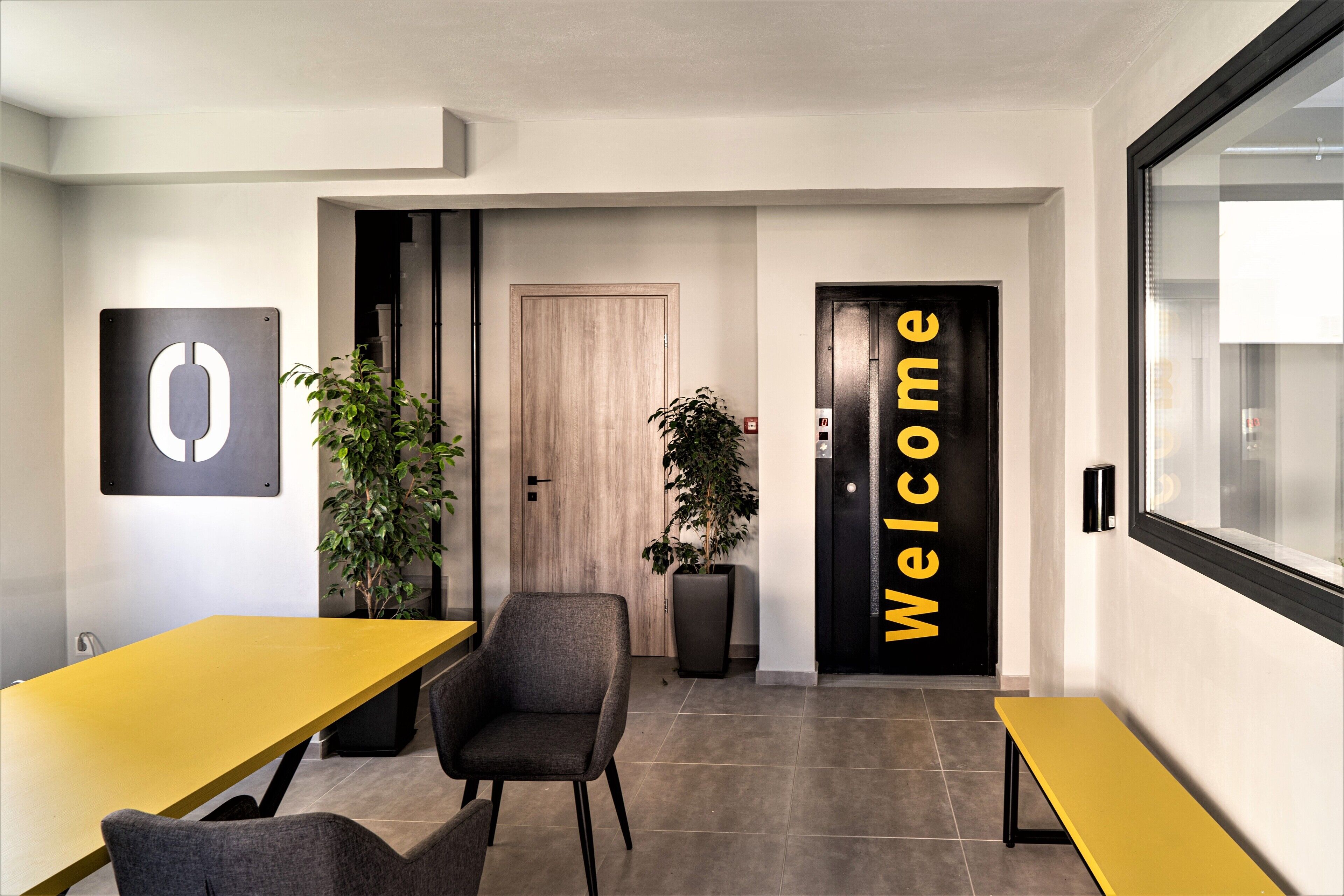 Ein moderner Arbeitsraum mit einem gelben Tisch, einem grauen Stuhl, einer großen Topfpflanze und einem Schild mit der Aufschrift 'Welcome' an der Tür.