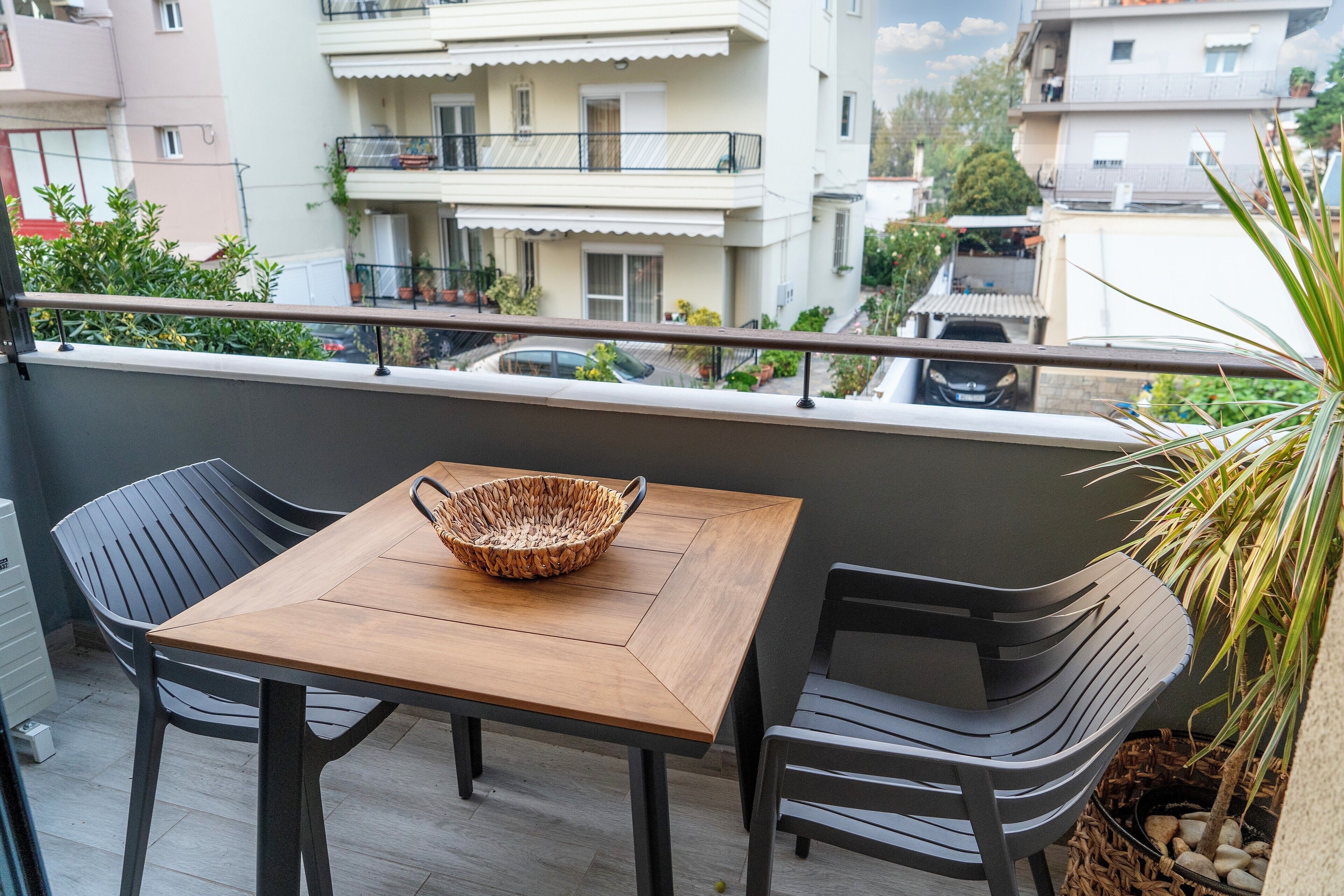 A balcony with a wooden table and two chairs, a woven basket on the table, and a potted plant.