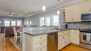 Cozinha moderna com armários brancos, bancada de granito, eletrodomésticos de aço inoxidável e área de jantar com sofá marrom e cadeiras brancas.