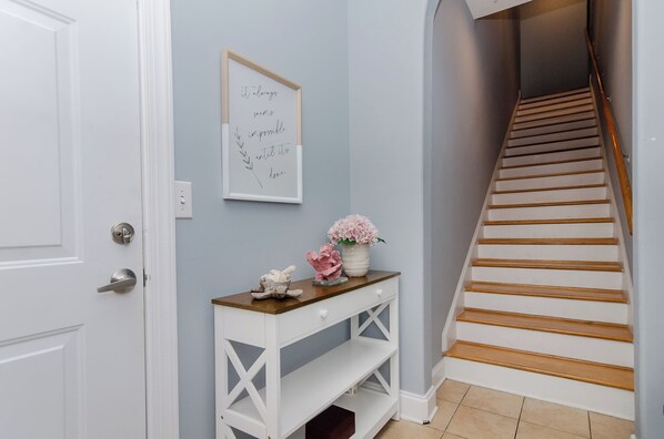 A hallway with a white door, a framed quote on the wall, a console table with a vase of flowers, and a wooden staircase.