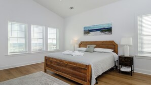 Quarto com cama de madeira, roupa de cama branca, pintura de uma praia e mesa de cabeceira com abajur.