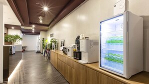 Un moderno vestíbulo de hotel con un mostrador de recepción de madera, un refrigerador grande repleto de bebidas y un espacio limpio, bien iluminado y con plantas en macetas.