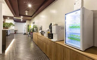 Um lobby de hotel moderno com uma mesa de recepção de madeira, uma geladeira grande abastecida com bebidas e um espaço limpo, bem iluminado e com plantas em vasos.