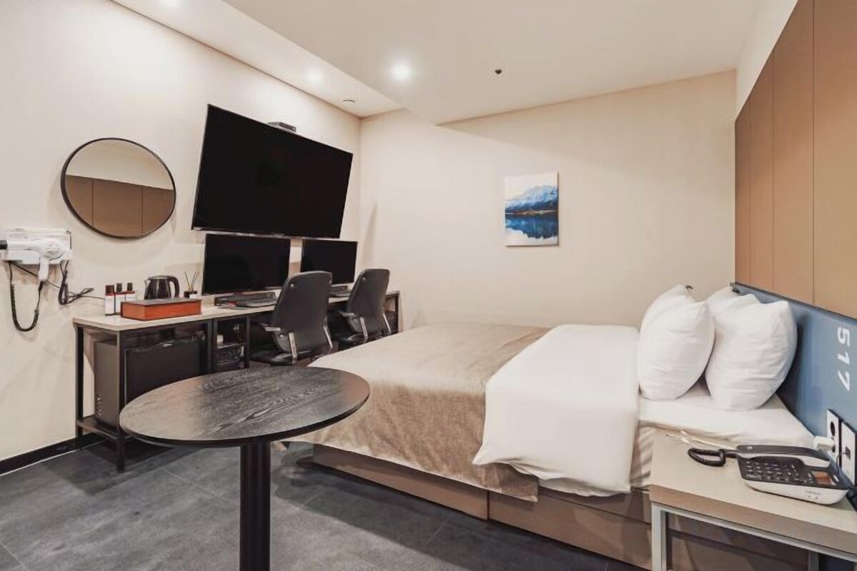A hotel room with a bed, a desk with a computer, a chair, a round table, a TV, and a painting on the wall.