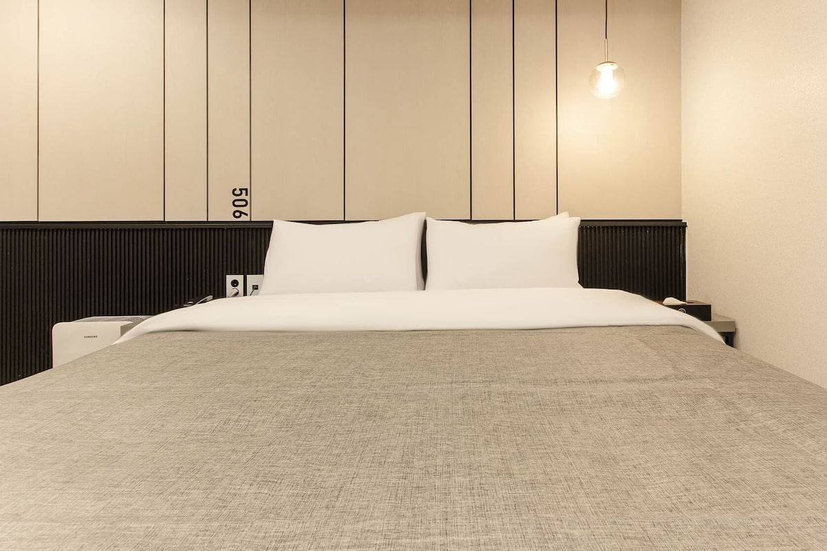 A neatly made bed with a grey blanket, a black headboard, and a single bedside lamp.