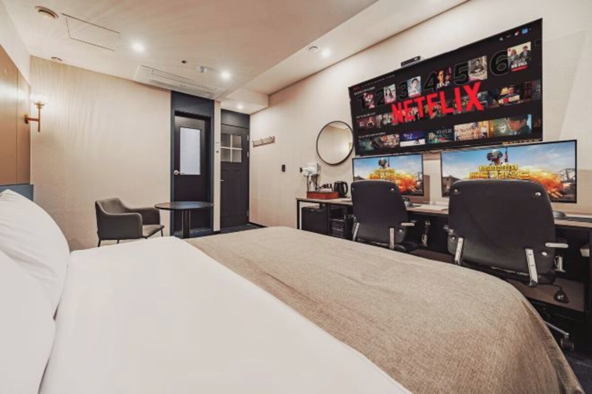 A hotel room with a bed, a desk with two chairs, a TV mounted on the wall, and a door.