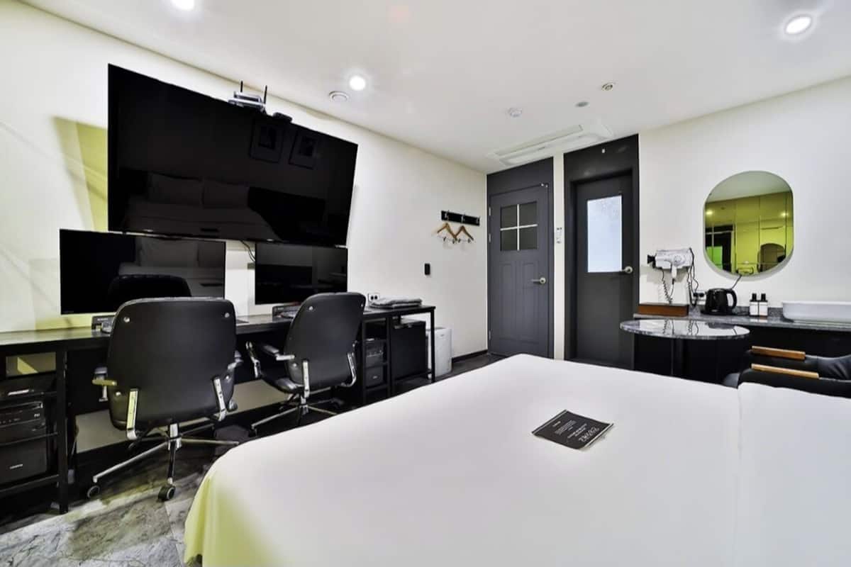 A modern hotel room with a large bed, a desk with two chairs, a kitchenette, a round mirror, and a door with a small window.
