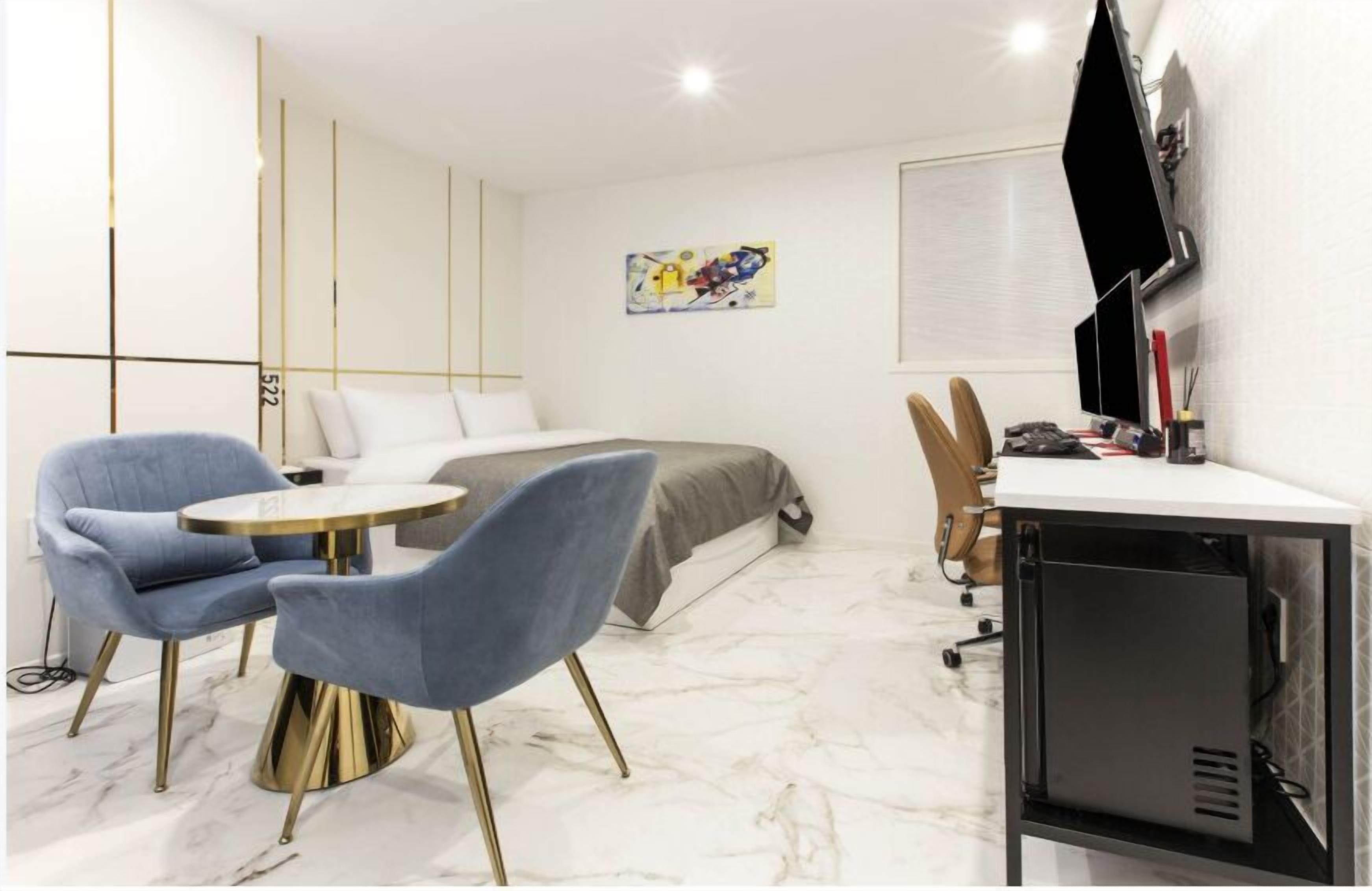 Quarto de hotel moderno com uma cama, uma mesa com computador, duas cadeiras azuis e uma pequena mesa.