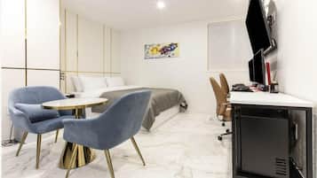 Quarto de hotel moderno com uma cama, uma mesa com computador, duas cadeiras azuis e uma pequena mesa.