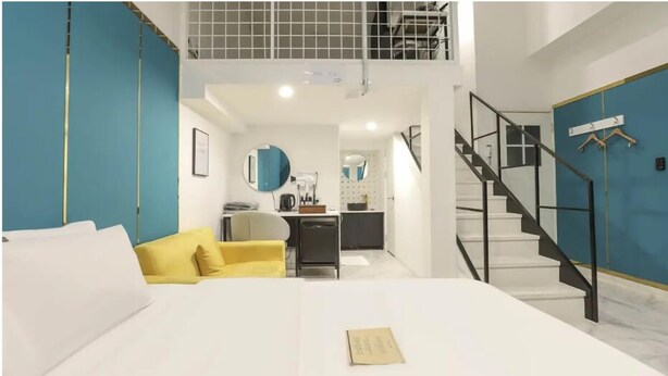Una habitación de hotel moderna con una cama, un sillón amarillo, una kitchenette y una escalera.