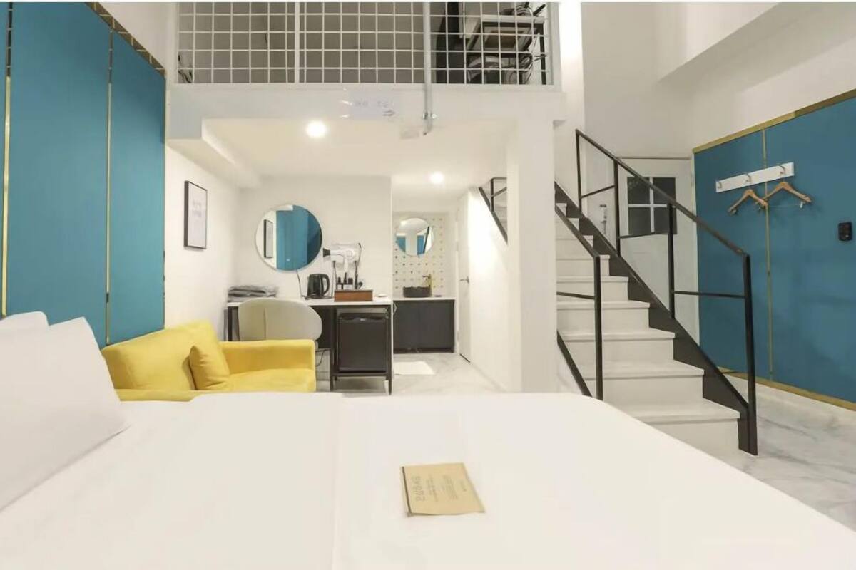 A modern hotel room with a bed, a yellow armchair, a kitchenette, and a staircase.