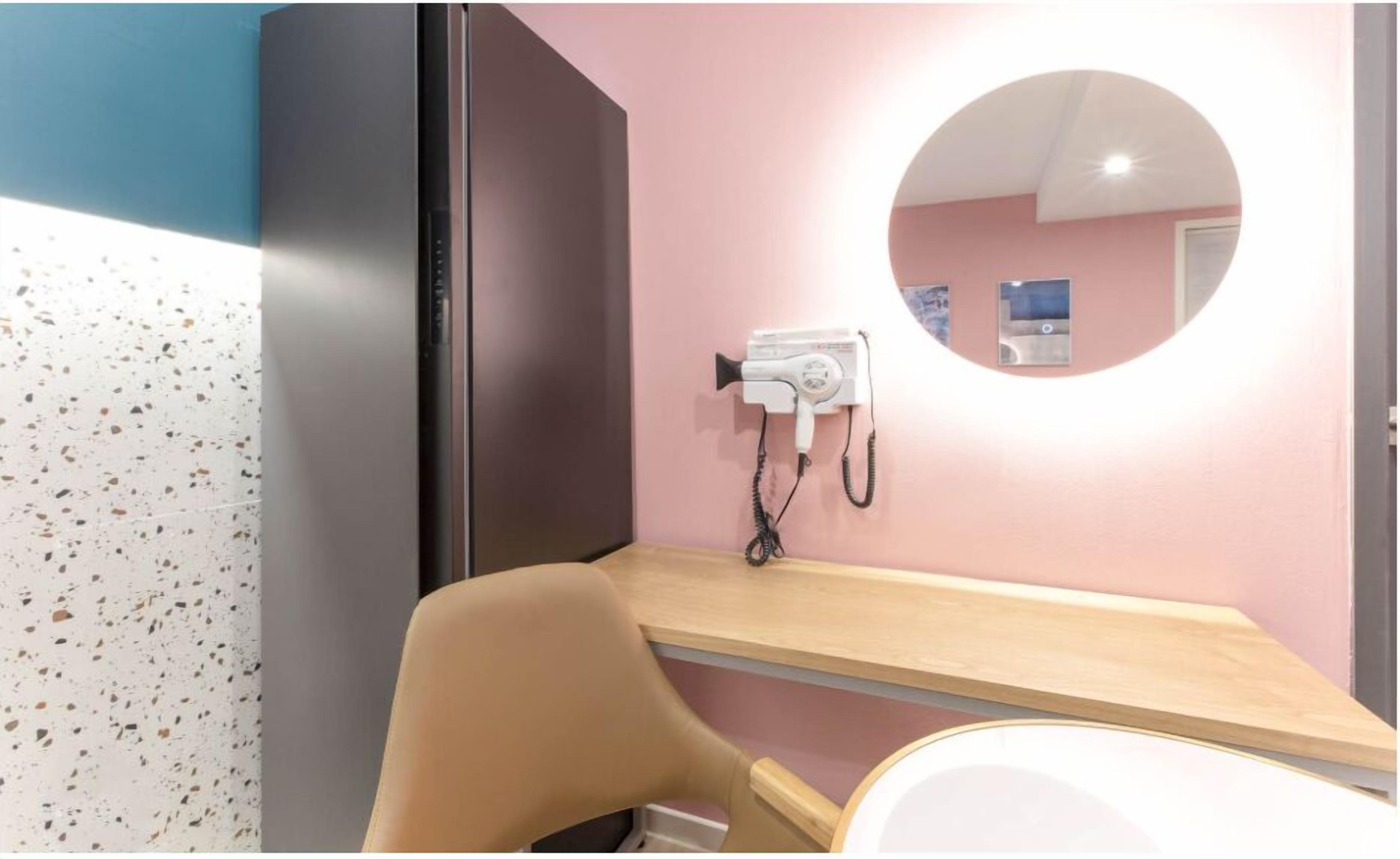Une salle de bain avec un mur rose, un miroir rond, un sèche-cheveux posé sur un support mural et un meuble-lavabo en bois.