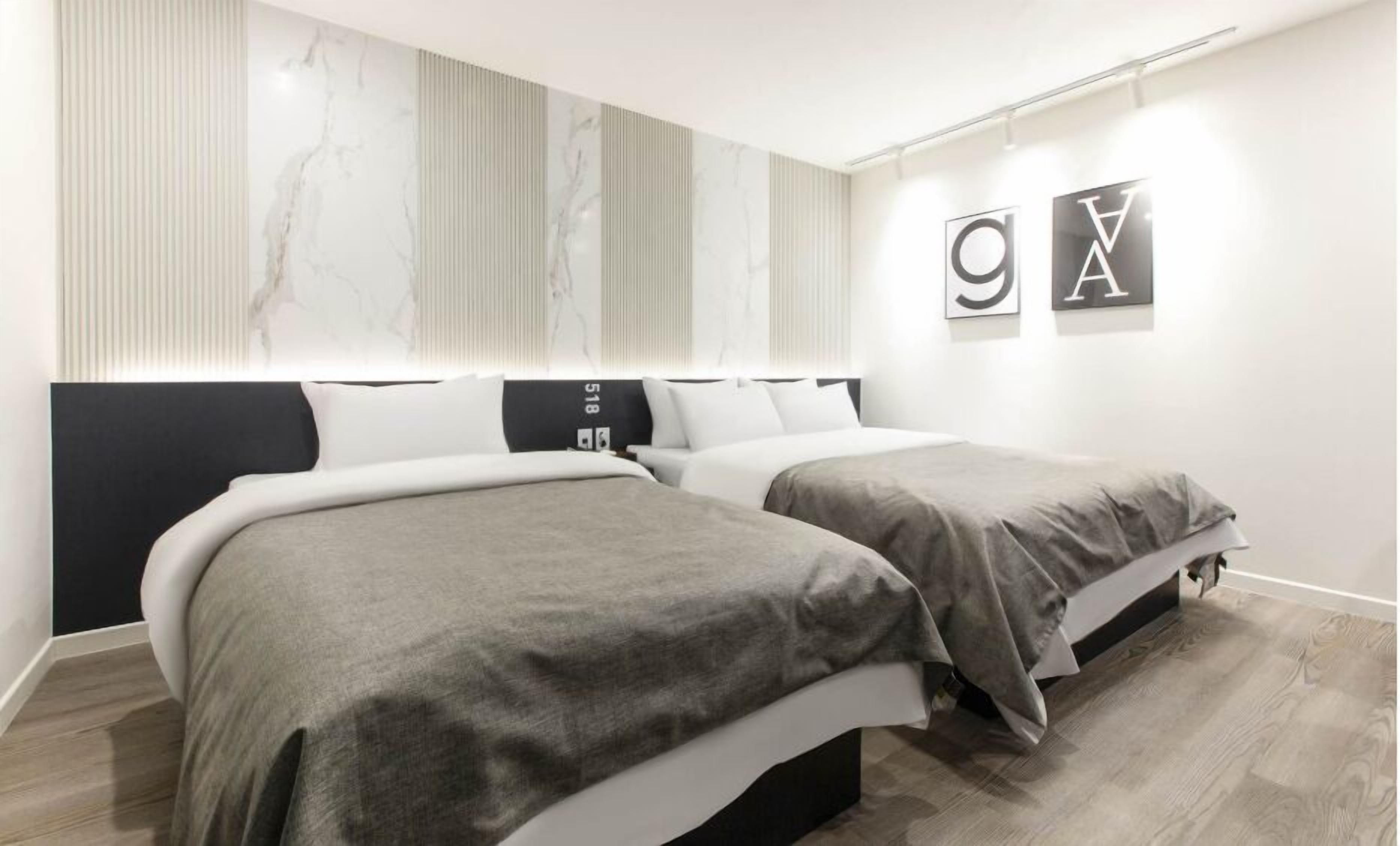 Una habitación de hotel moderna con dos camas, un ventanal con persianas y una pared con arte abstracto y las letras 'GVA'.