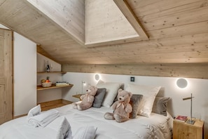 A bedroom with a wooden ceiling, a bed with white bedding, two teddy bears, a bedside table with a book and a glass, and a shelf with a basket of fruit and a wine bottle.