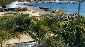 Una marina costera con barcos amarrados, una playa arenosa y colinas y edificios al fondo.