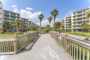 A wooden boardwalk leading to a hotel with multiple balconies and palm trees.