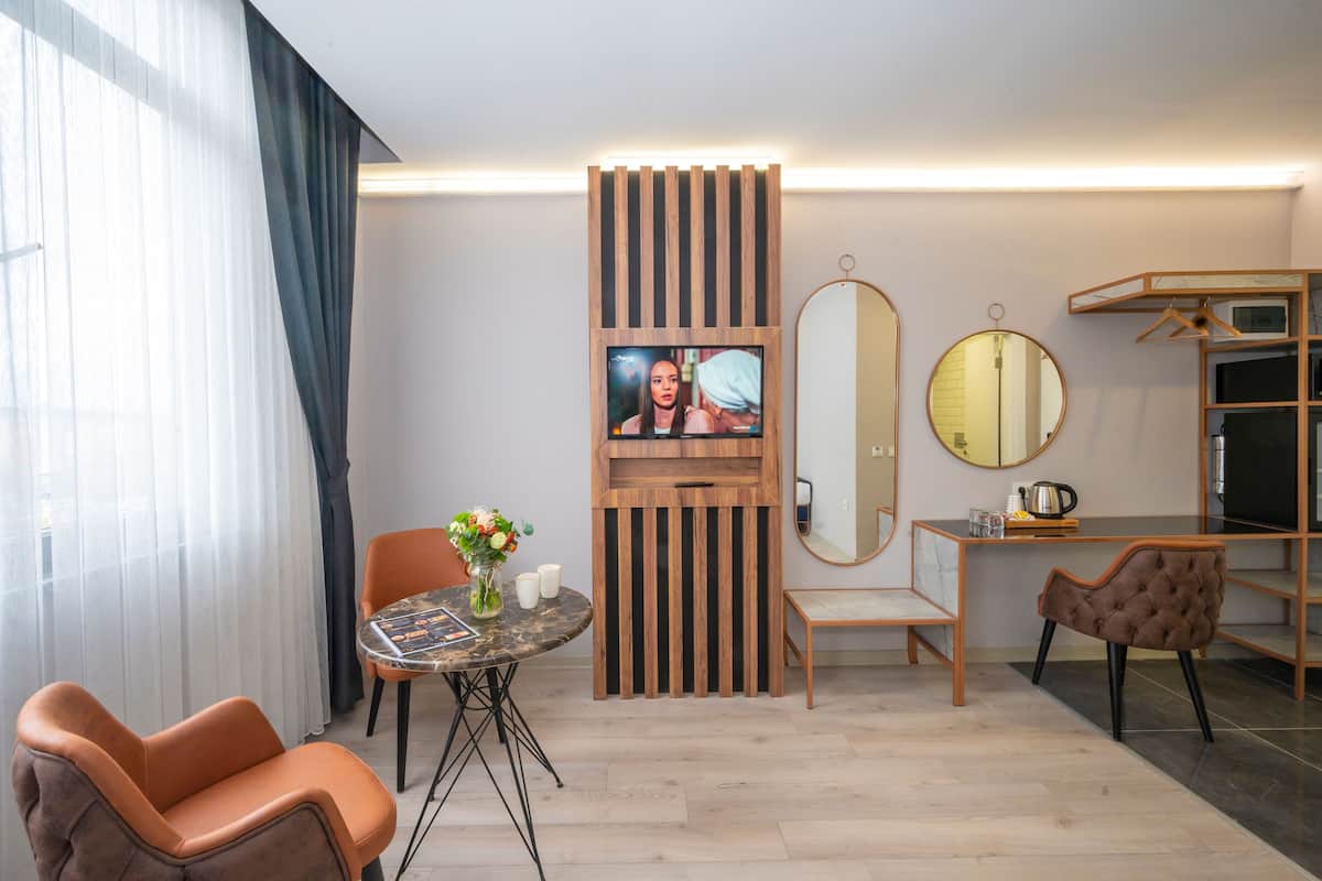 A modern hotel room with a large window, a wooden accent wall, a round mirror, a TV, a dining table with chairs, and a sofa.