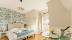 Quarto com cama, mesa de cabeceira, janela com cortinas, mesa de jantar com uma cadeira e uma garrafa de champanhe.