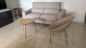 Ett vardagsrum med en soffa, ett litet runt bord, en flätstol, en tv-apparat på ett skåp och en tavla på väggen.