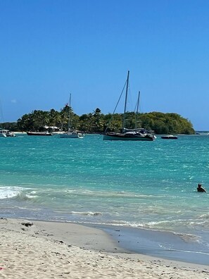 A beach with clear turquoise water, a sandy shore, and several sailboats anchored near a small island with dense vegetation.
